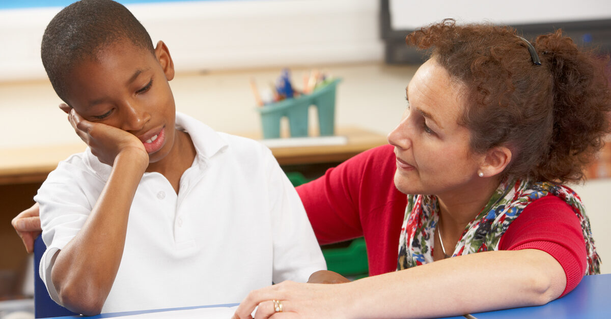 In a Looped classroom, boy looks unhappy as teacher tries to explain something at his desk.